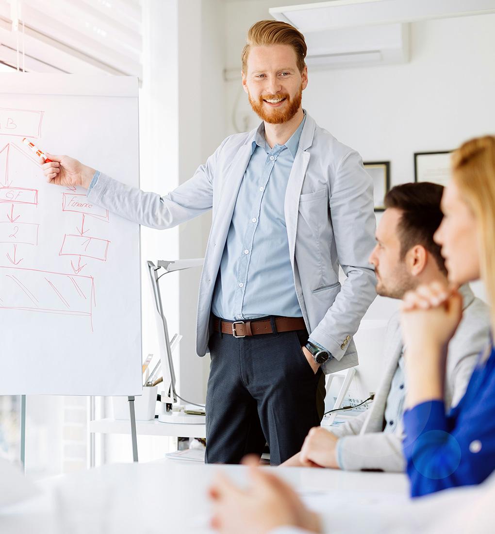 A smiling consultant stood pointing at a diagram covered whiteboard with a pen while giving a presentation to clients in a well lit, professional looking office.