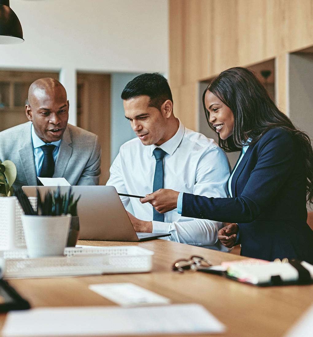 A group of professionals in suits sat around a table looking at a laptop discussing discussing work while smiling.