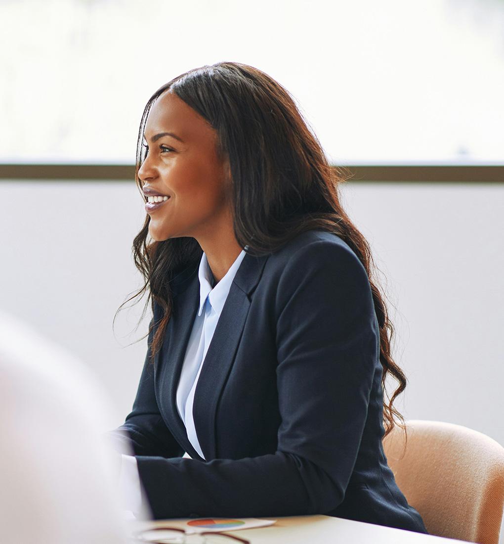 A smiling professional wearing a suit sat at a table in an interview while looking engaged.