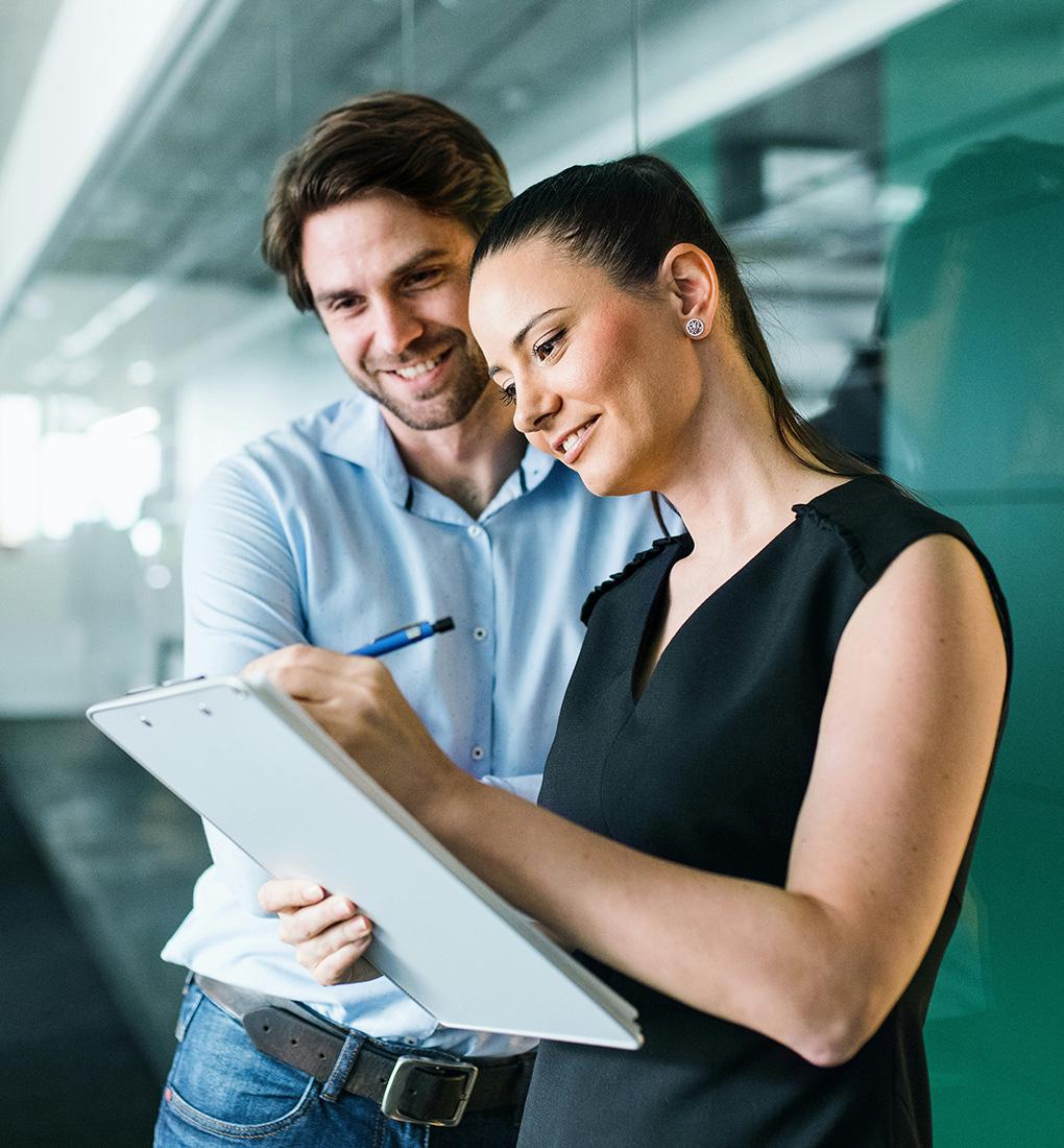 Two professionals in an office looking intently at a clipboard and taking notes while smiling.