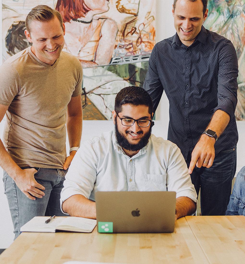 A man sat working at a laptop in an office with his two coworkers behind him looking at is work while smiling.